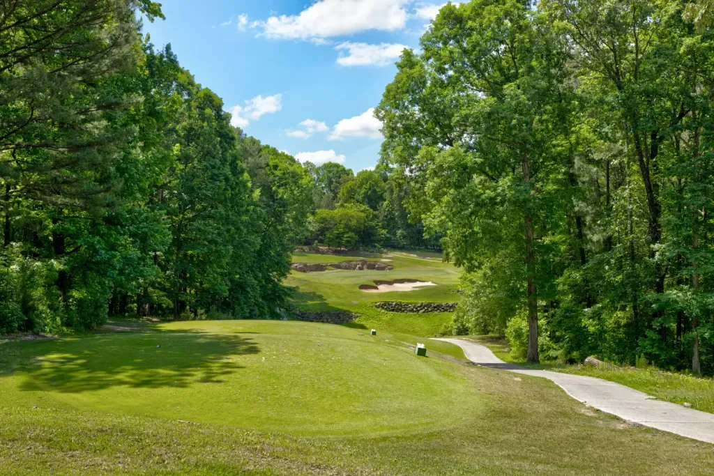 A scenic golf course with a green fairway, trees on both sides, and sand bunkers in the distance under a partly cloudy sky.
