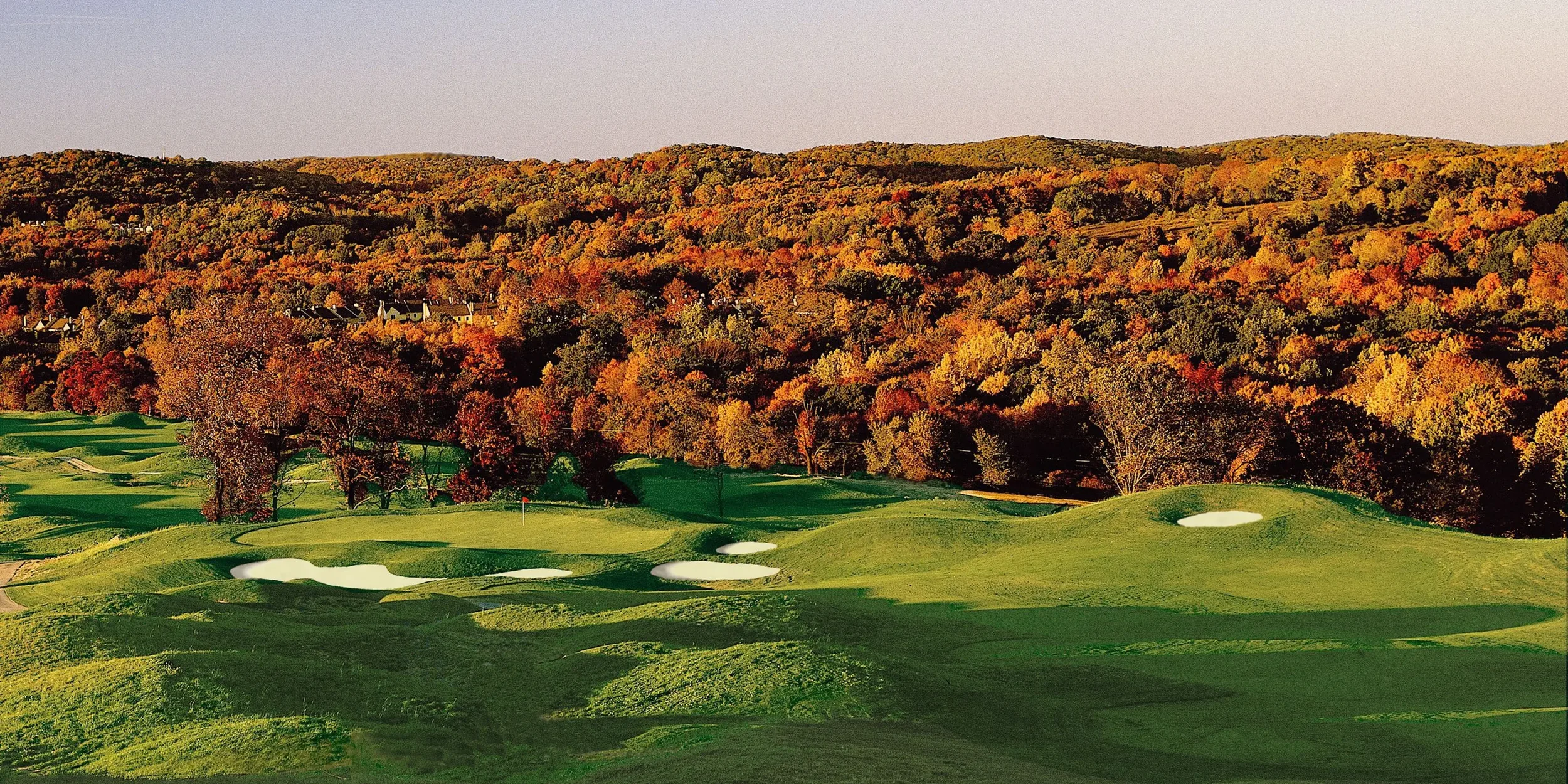 A golf course with green fairways and sand bunkers, set against a background of colorful autumn trees on rolling hills.