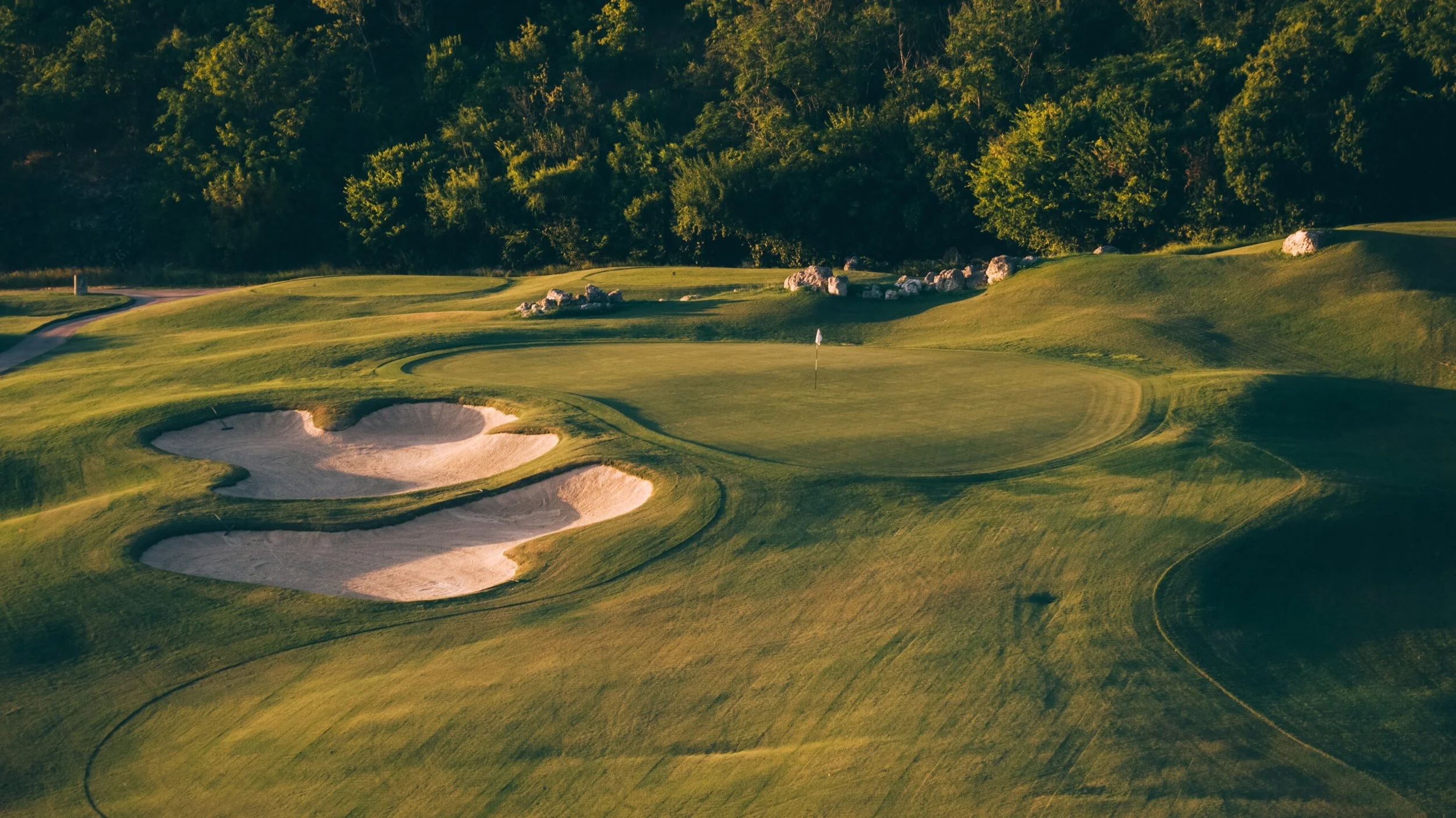 A golf course with green grass, sand bunkers, a flagstick on the putting green, and trees in the background.