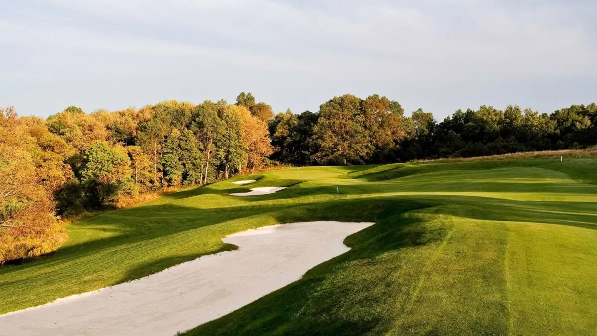 A golf course with sand traps and green fairways, surrounded by trees with autumn foliage under a partly cloudy sky.