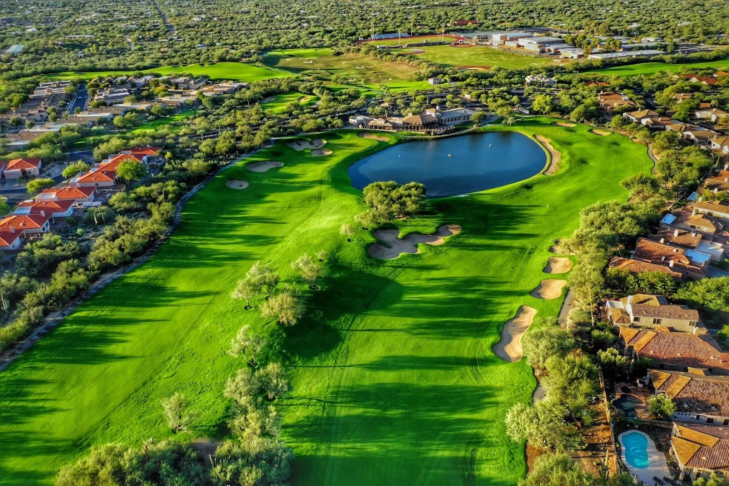 Aerial view of a golf course with green fairways, sand traps, a water hazard, surrounded by residential houses and trees.