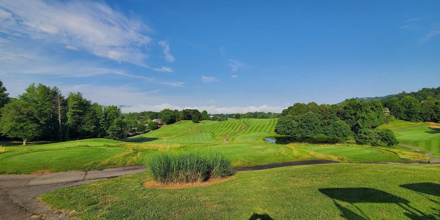 A scenic view of a lush green golf course with rolling hills, trees, a small pond, and a clear blue sky.