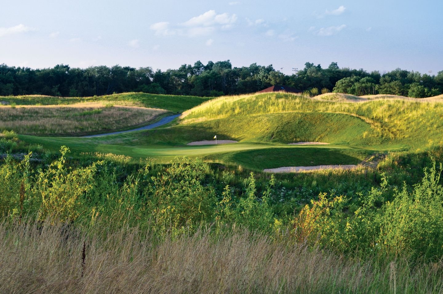 A scenic golf course with a flag on the putting green surrounded by sand traps, rolling green hills, and trees in the background under a partly cloudy sky.