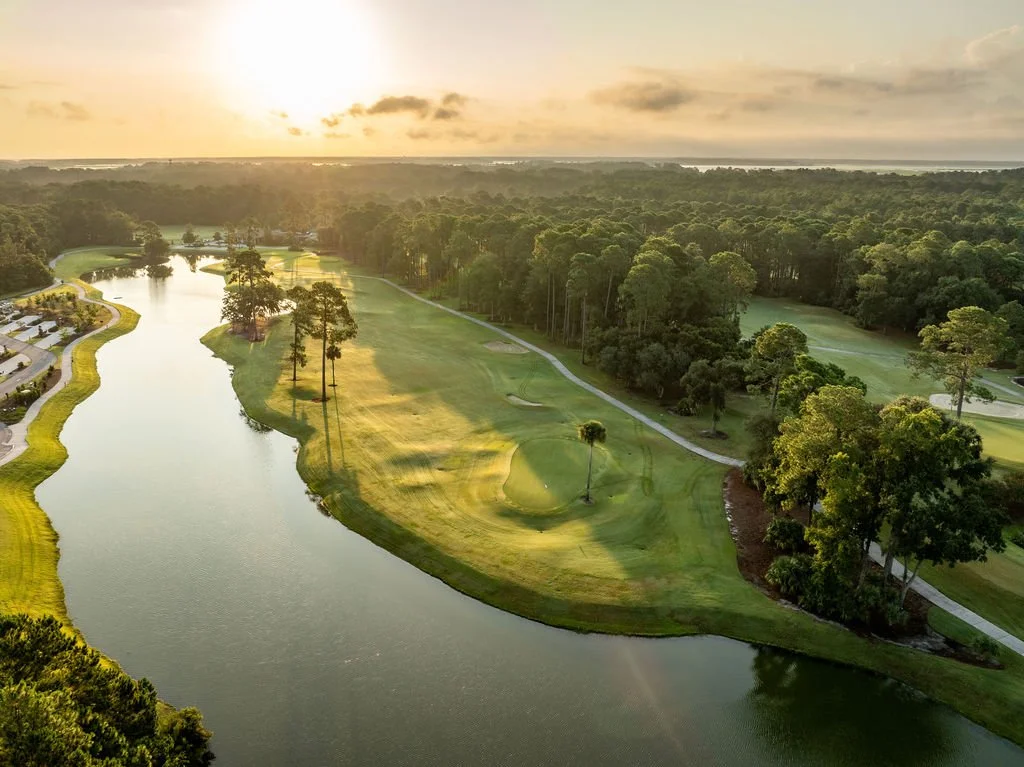 Aerial view of a golf course at sunset with a water hazard, grass fairways, and trees.