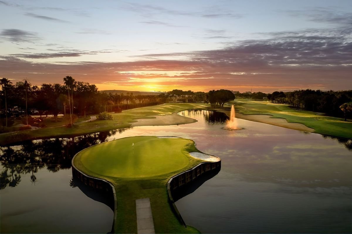 A golf course with a water hazard and fountain, trees lining the course, and a sunset sky in the background.