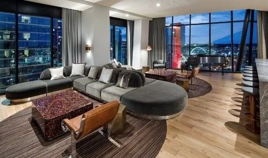 Modern living room with large windows, city skyline view, gray sectional sofa, coffee tables, wooden flooring, and a grand piano.
