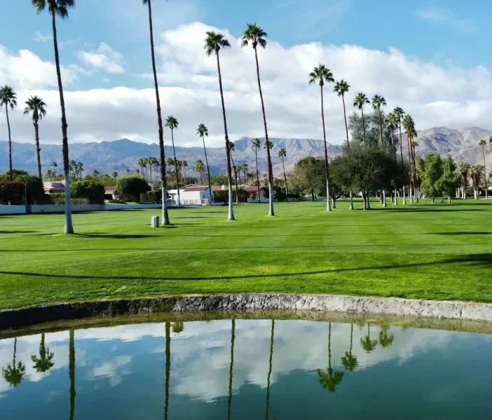 A golf course with tall palm trees, green grass, a water hazard reflecting the sky, and mountains in the background under a partly cloudy sky.