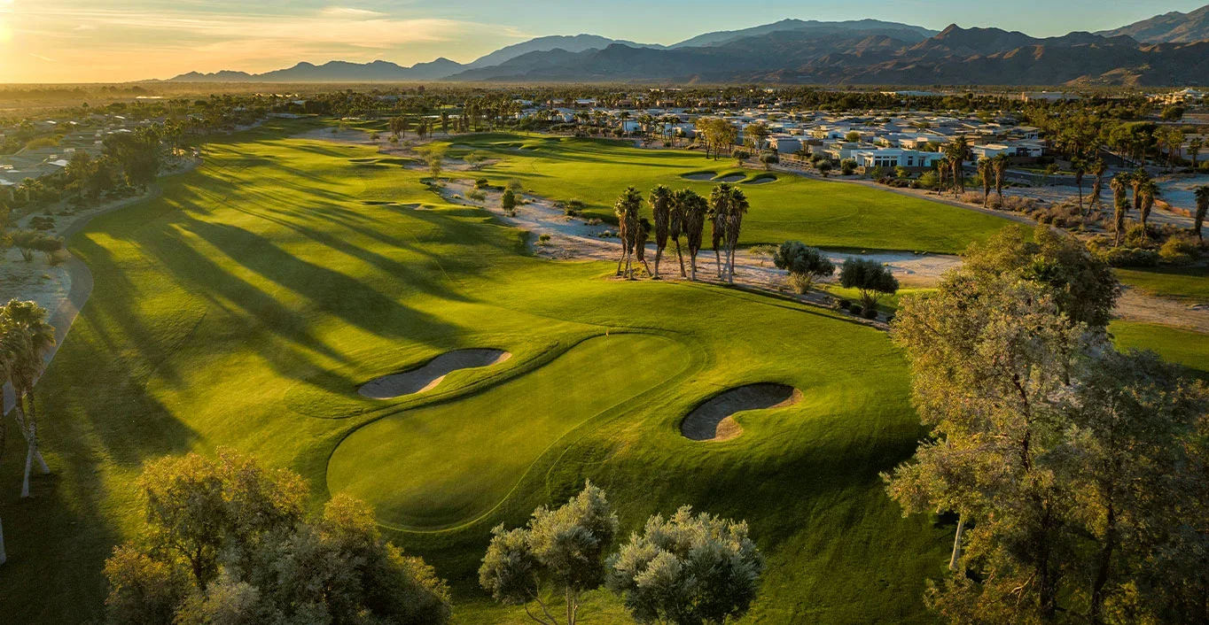 Aerial view of a lush golf course at sunset, surrounded by trees, with mountains in the background.