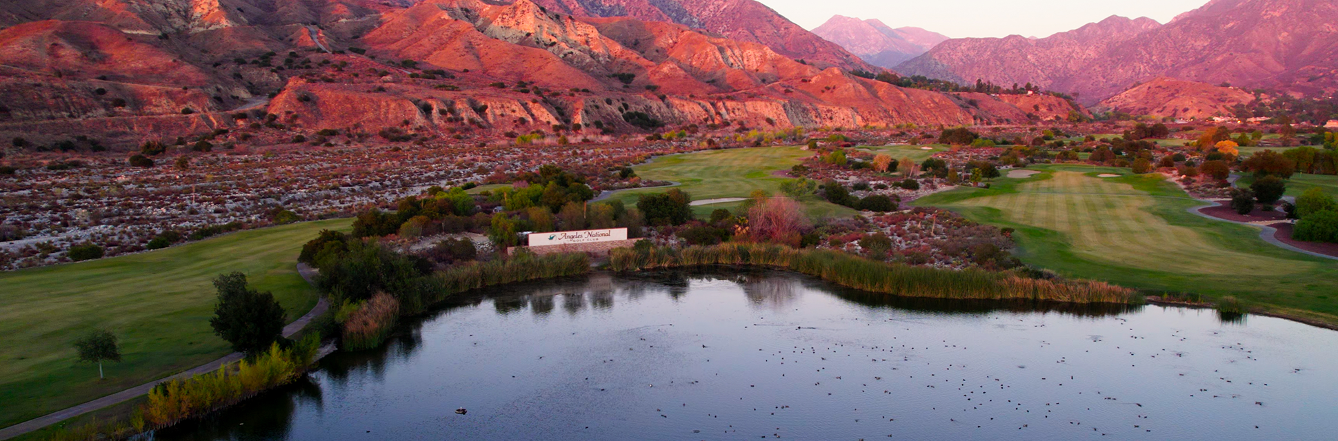 A scenic landscape of a golf course with green fairways, a pond in the foreground, and rolling hills or mountains in the background at sunset.