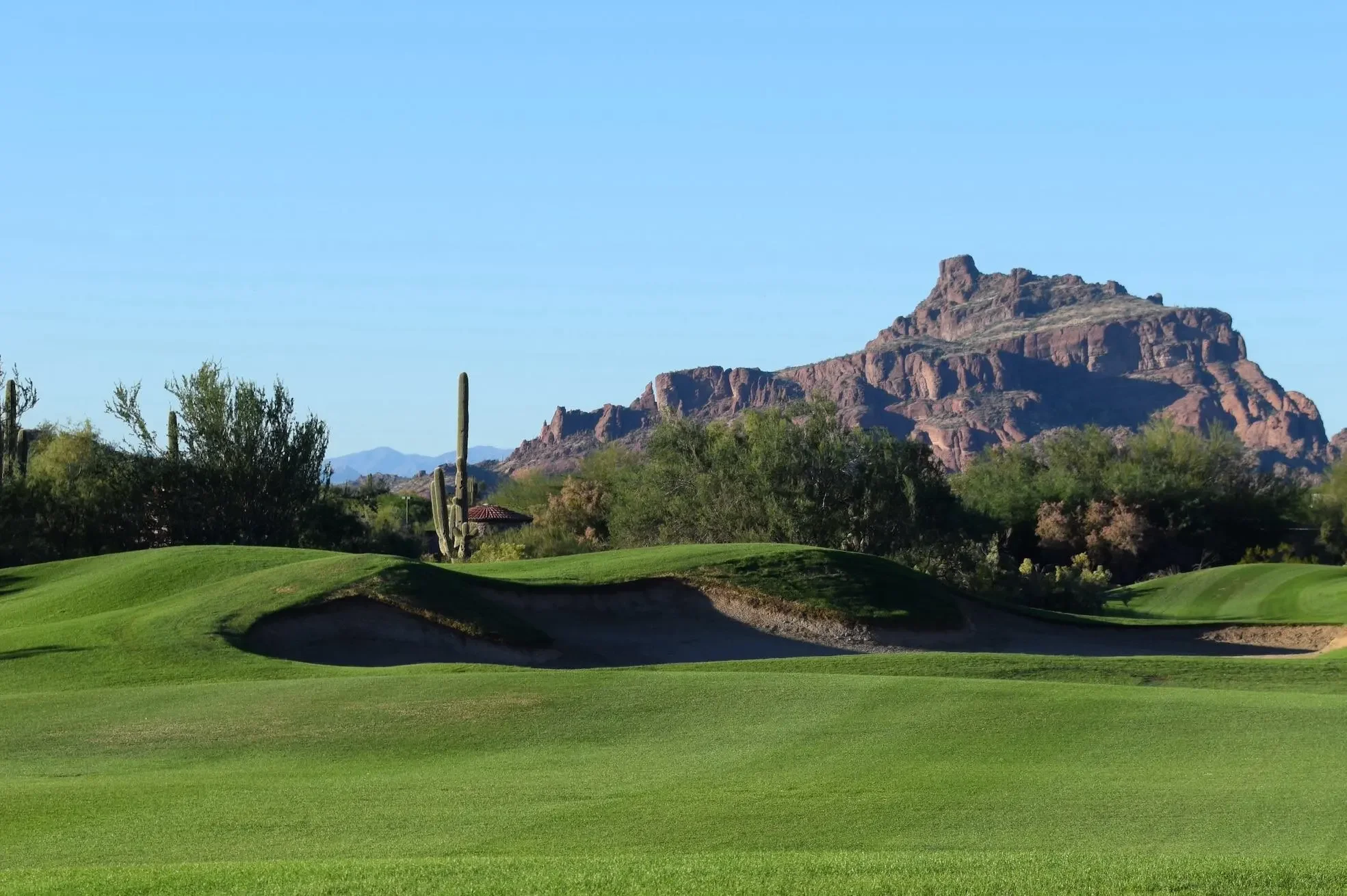 A scenic golf course with a green lawn and sand bunkers, desert vegetation, cacti, and a mountain in the background under a clear blue sky.