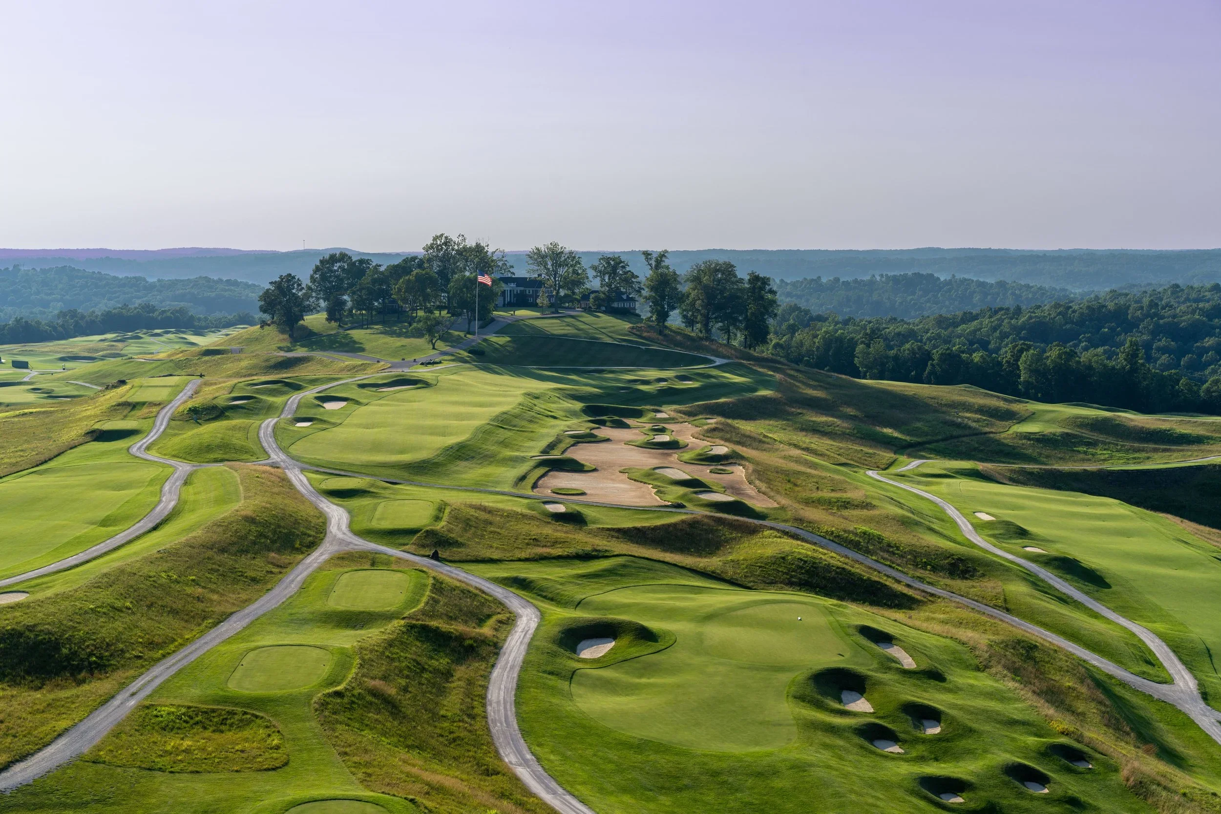 An aerial view of a golf course on a hilly landscape with multiple greens, sand traps, and winding golf cart paths, surrounded by trees and distant hills.