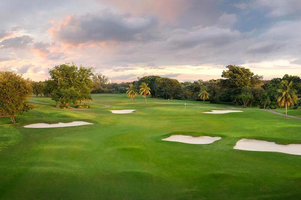 Scenic golf course with sand bunkers, green grass, trees, and cloudy sky at sunset.