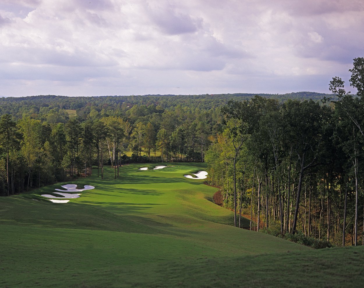 A scenic golf course with lush green fairways, sand traps, and a wooded background under a partly cloudy sky.