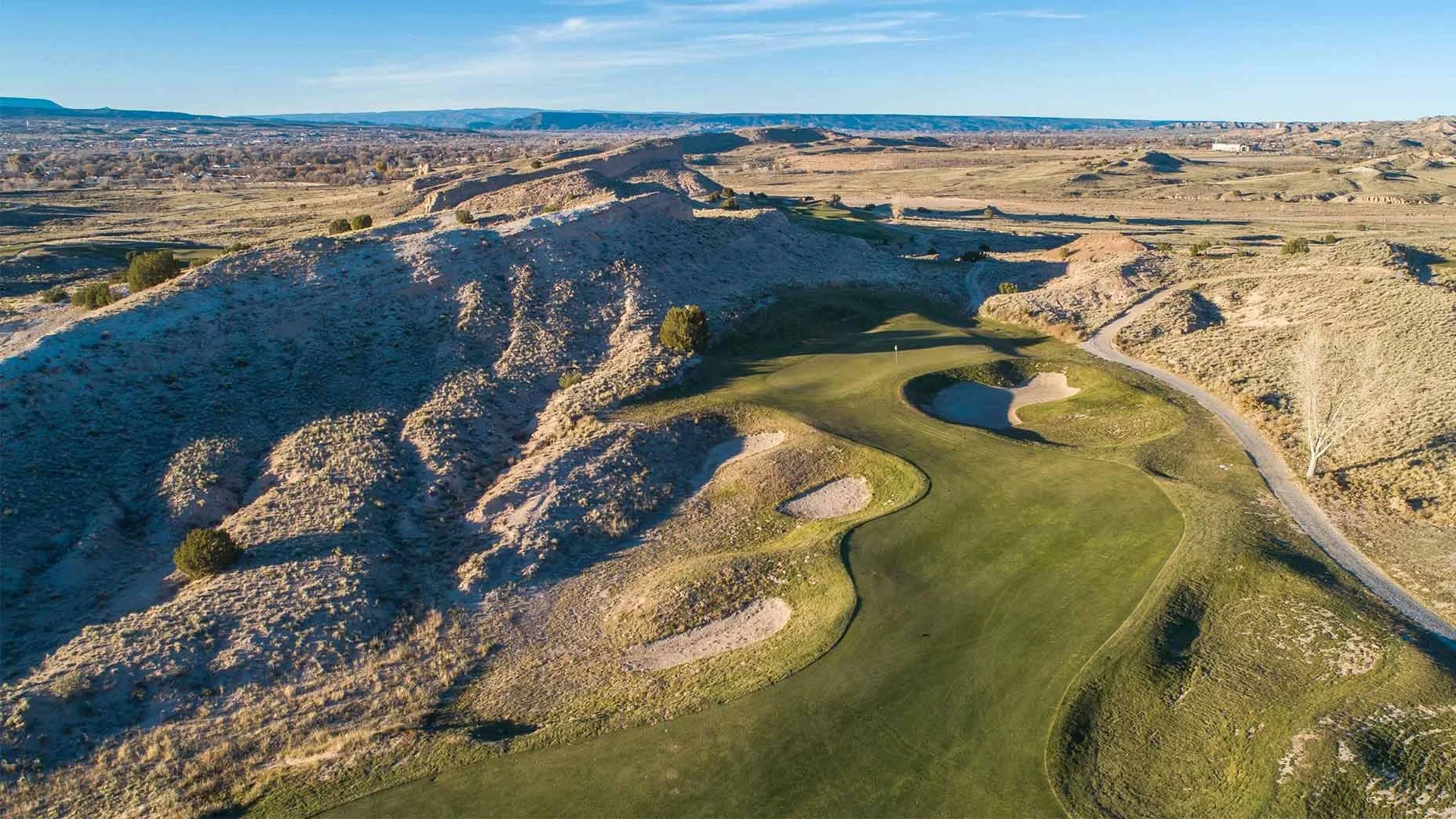 Aerial view of a golf course set in a hilly, desert-like landscape with sandy hills, sparse vegetation, and a small pond near the green, under a clear blue sky.