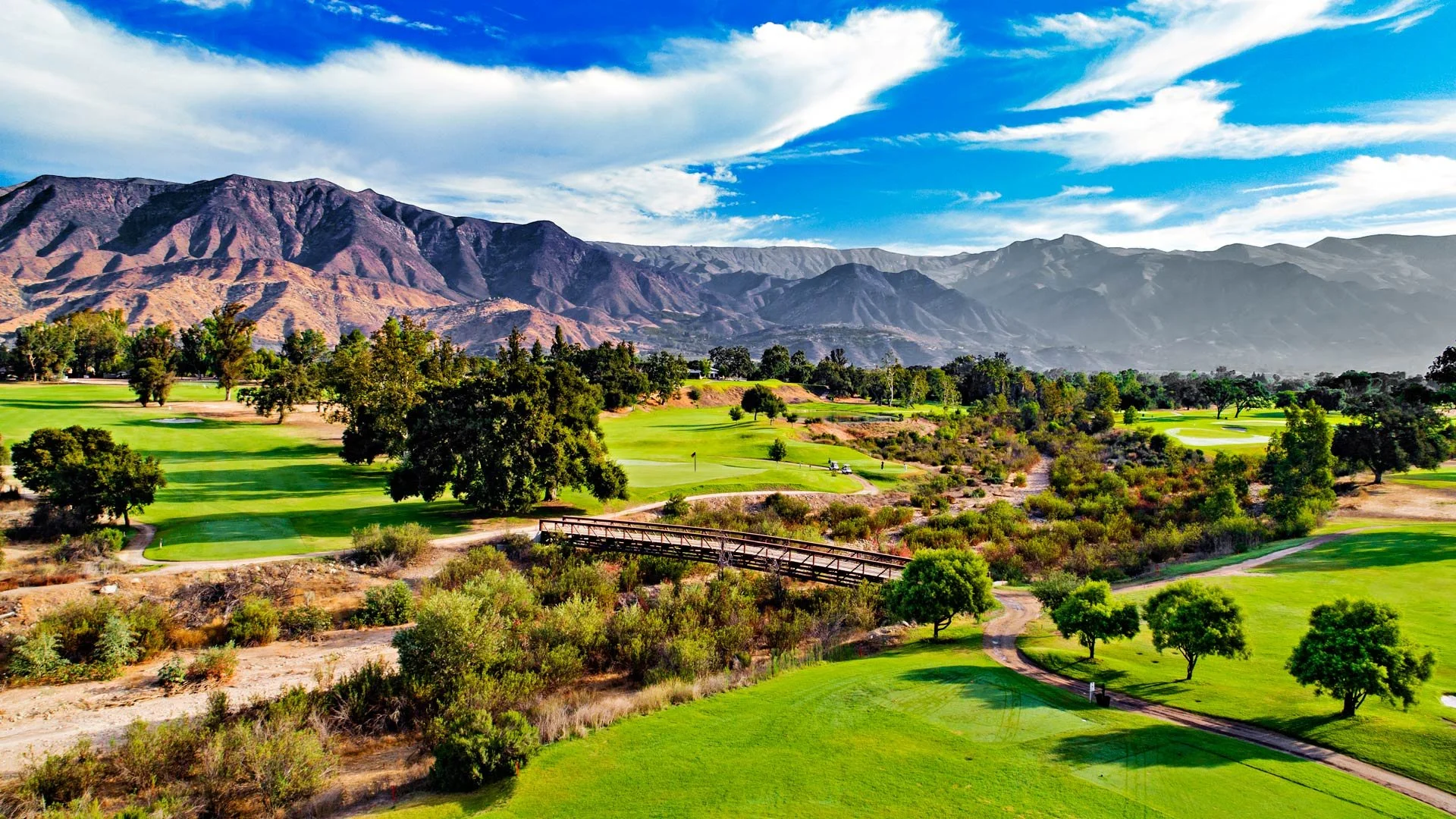 A scenic golf course surrounded by trees, with green fairways, a small wooden bridge, and mountains in the background under a partly cloudy sky.