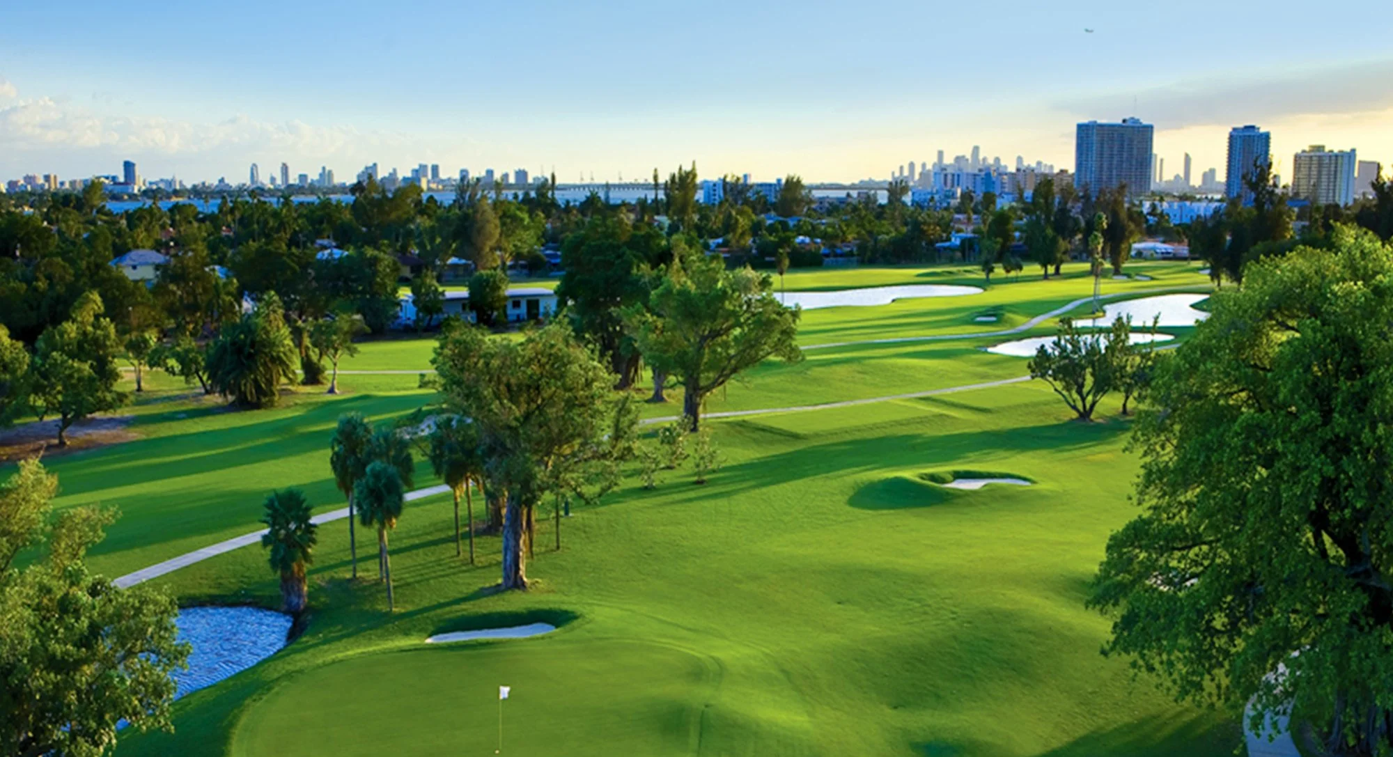 View of a golf course with green grass, sand traps, trees, and a city skyline in the background under a partly cloudy sky.