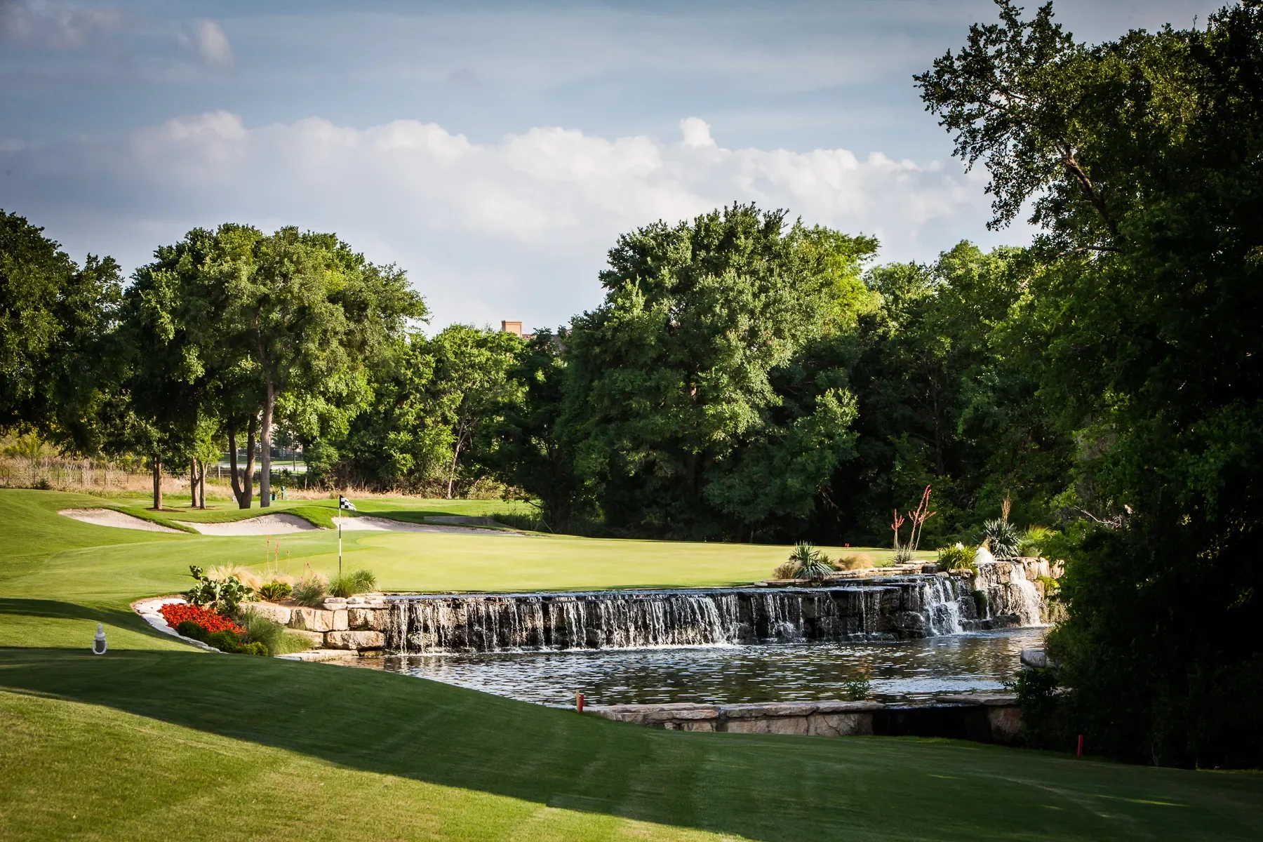 Golf course with a small waterfall and pond, green grass fairway, sand traps, a golf hole flag, and trees in the background under a cloudy sky.