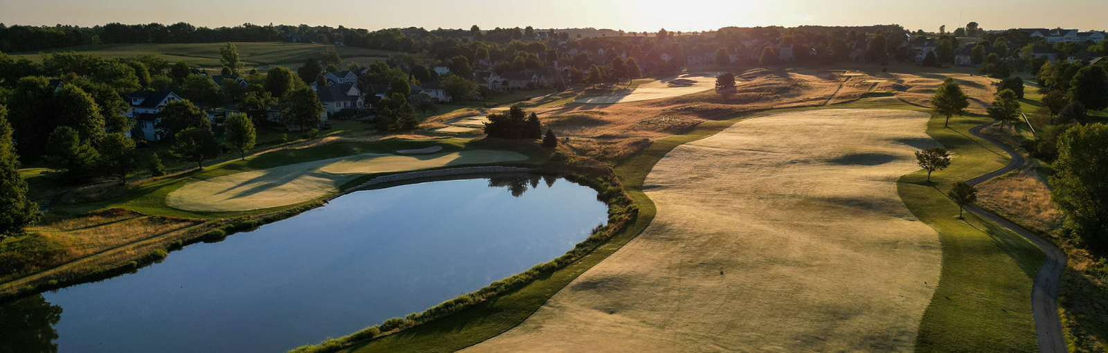 Aerial view of a golf course with a large pond, green fairways, and scattered trees, with houses in the background at sunset.
