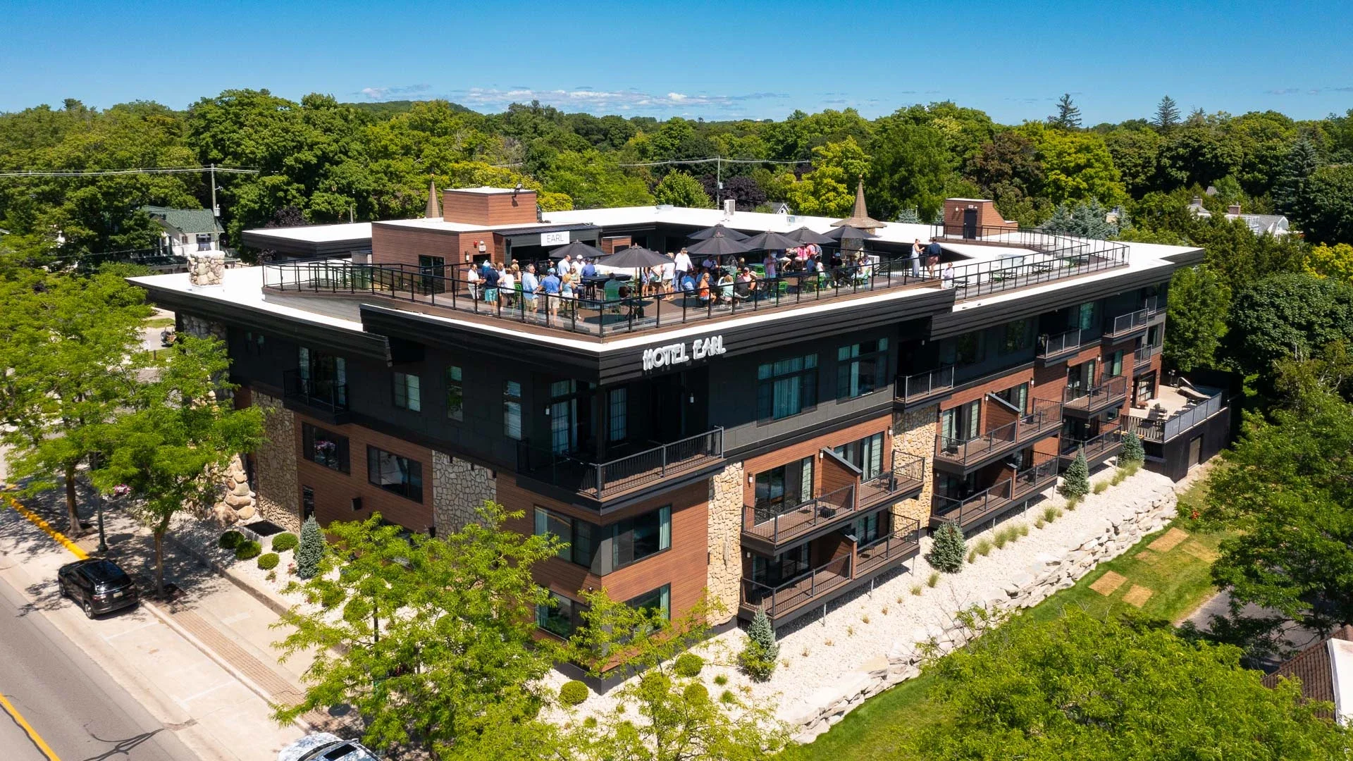 Aerial view of a modern hotel named 'Hotel Earl' with a rooftop terrace filled with people, umbrellas, and outdoor seating, surrounded by lush green trees and a clear blue sky.