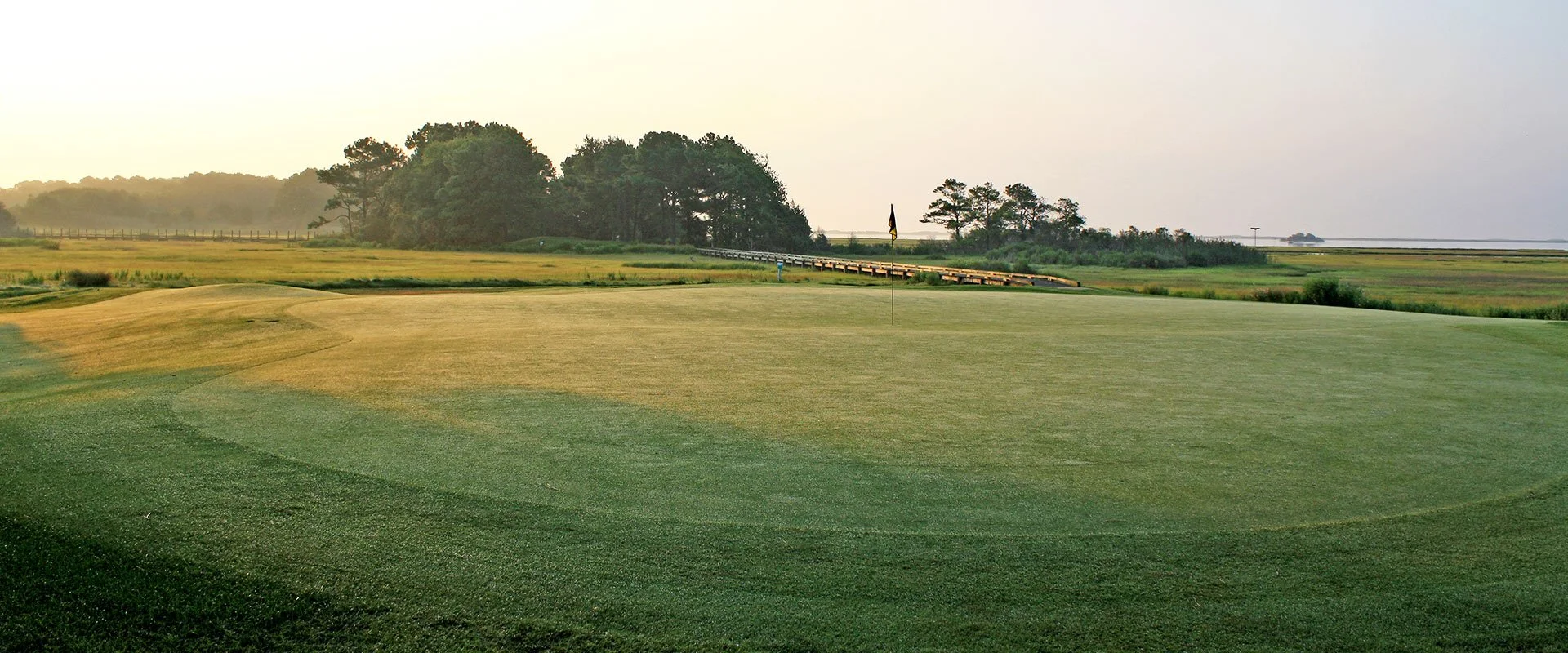 A golf course at sunrise with a flag on the green, trees in the background, and a misty sky.