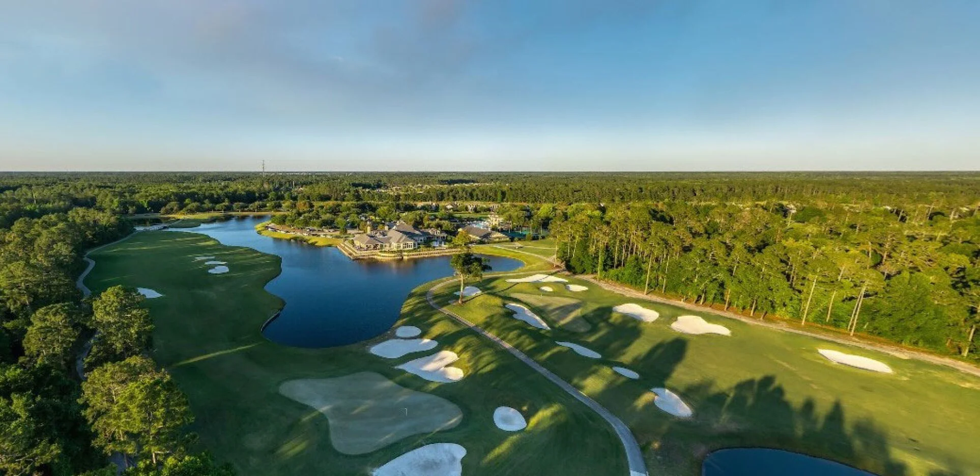 Aerial view of a golf course with green fairways, sand bunkers, water hazards, surrounded by trees, and a clubhouse in the background under a clear blue sky.