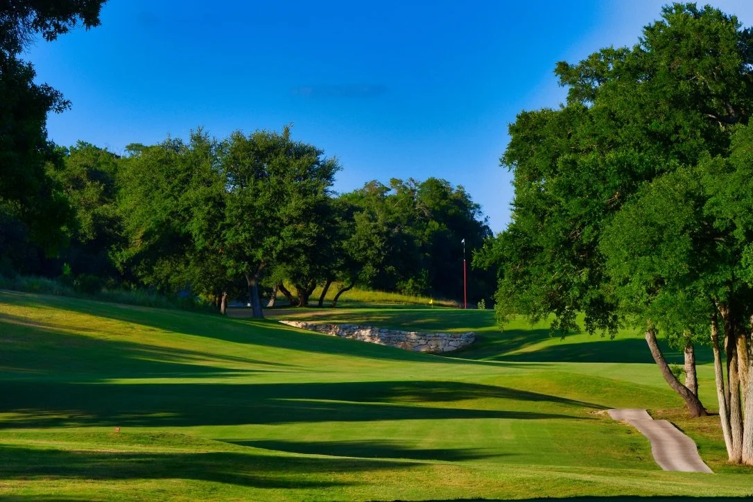 A lush green golf course with trees lining the fairway, a sand bunker, and a flag marking the hole in the distance under a clear blue sky.