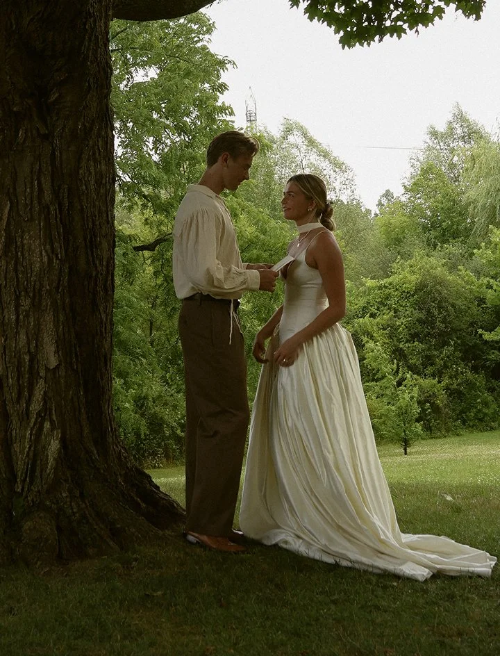 A man and woman dressed in vintage wedding attire standing outdoors next to a large tree, looking into each other's eyes during a wedding or romantic moment.