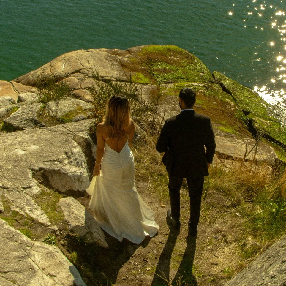 Bride and groom walking toward the water over sunlit rocks, photographed in a cinematic, documentary style by Sela Volk.