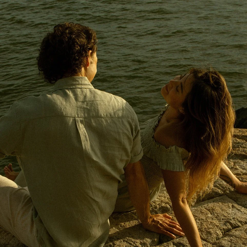 A man and woman sitting on rocks by the water, facing each other. The woman has long hair and is leaning back, while the man has curly hair and is sitting with his back to the camera, looking at the woman.