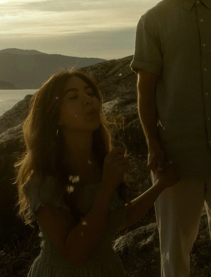 A woman with long hair holding hands with a man outdoors near a rocky shoreline during sunset, with mountains and water visible in the background.