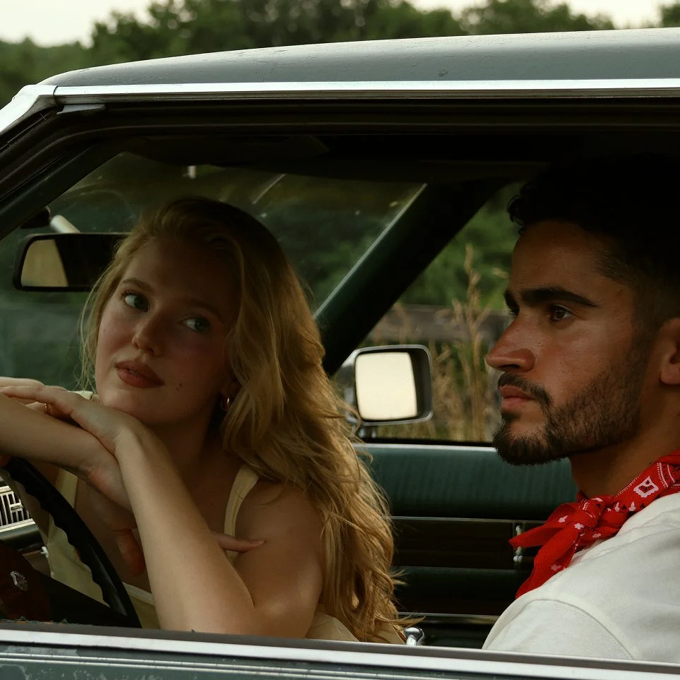 A young woman with blonde hair and a young man with dark hair and a red bandana sitting inside a vintage car, both looking seriously ahead.