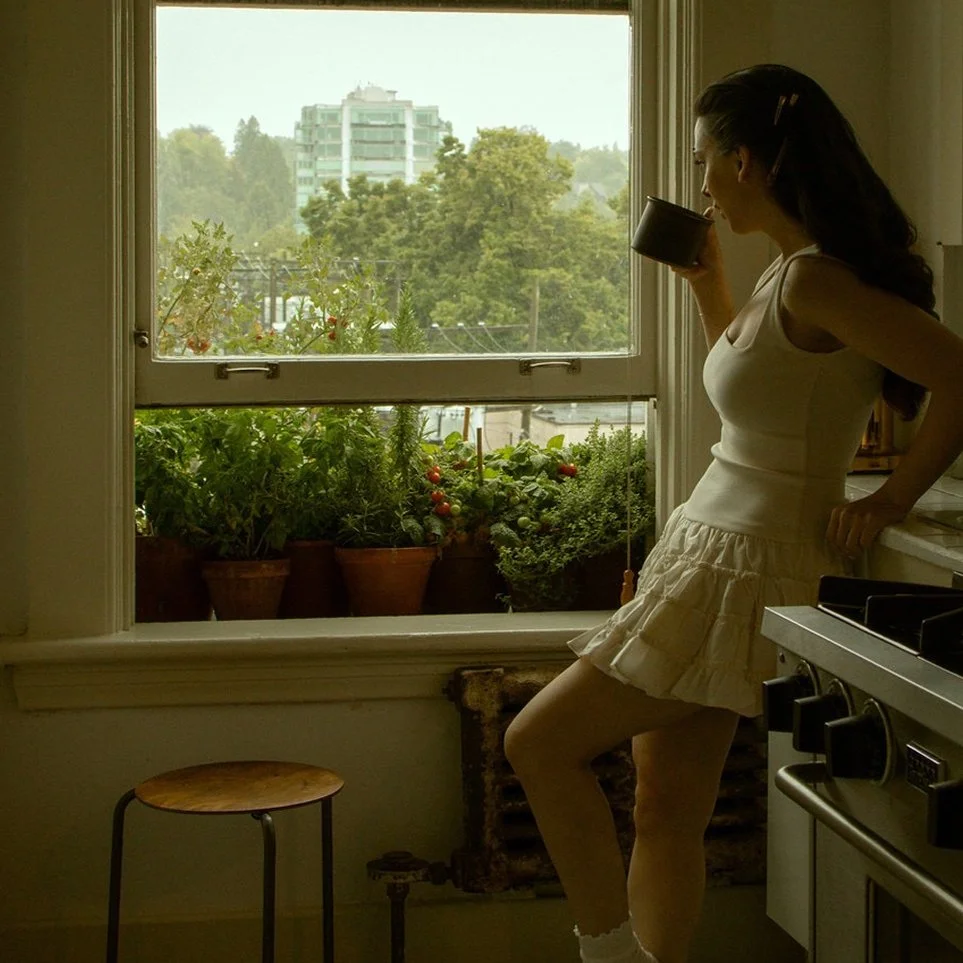 A woman standing in a kitchen near a window, drinking from a cup, with a view of potted plants and a city skyline outside.