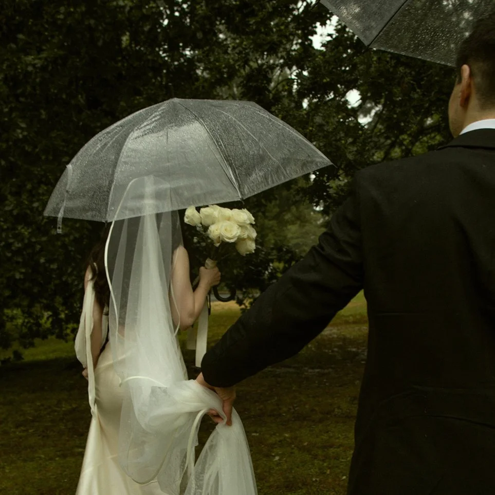 Bride and groom walking under clear umbrellas in the rain, captured in a cinematic, storytelling wedding photograph by Sela Volk.