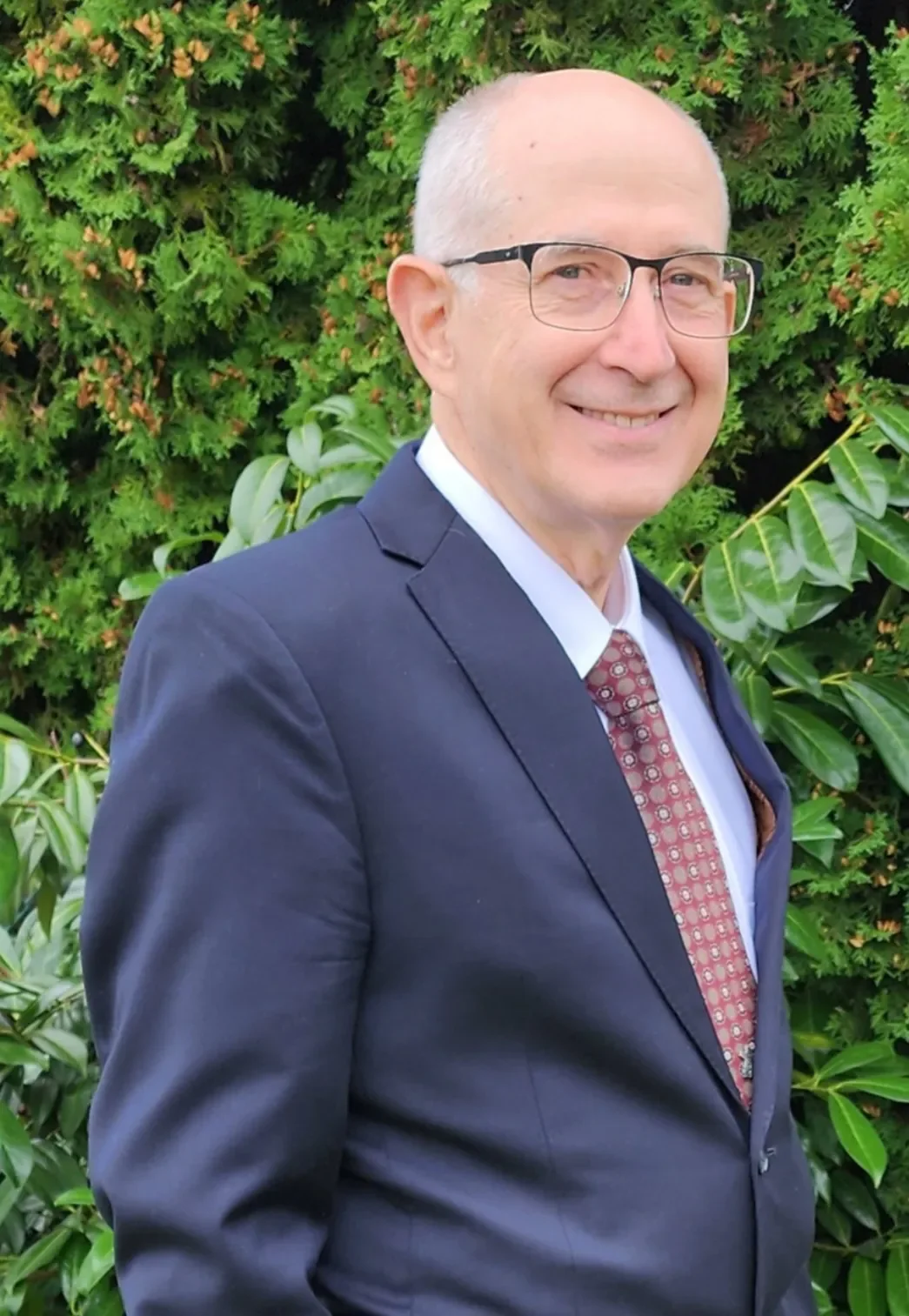 A middle-aged man with glasses, wearing a dark suit, white shirt, and patterned tie, smiling outdoors in front of green foliage.