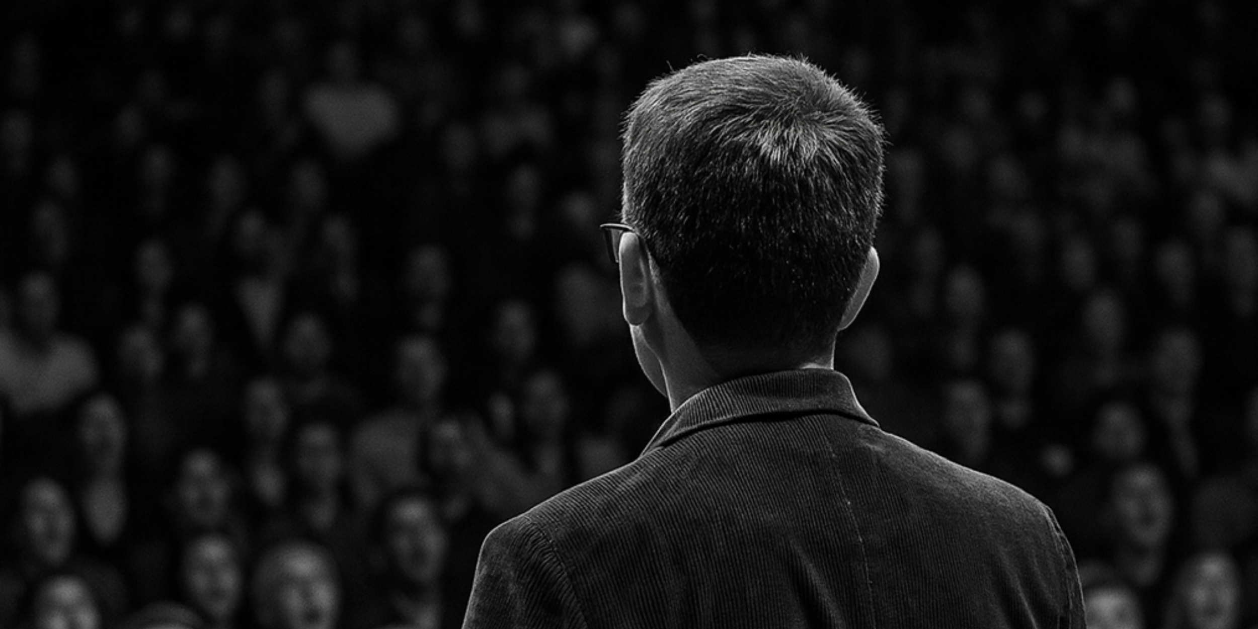 A man with short hair and glasses is seen from behind addressing a large audience in a dark setting.