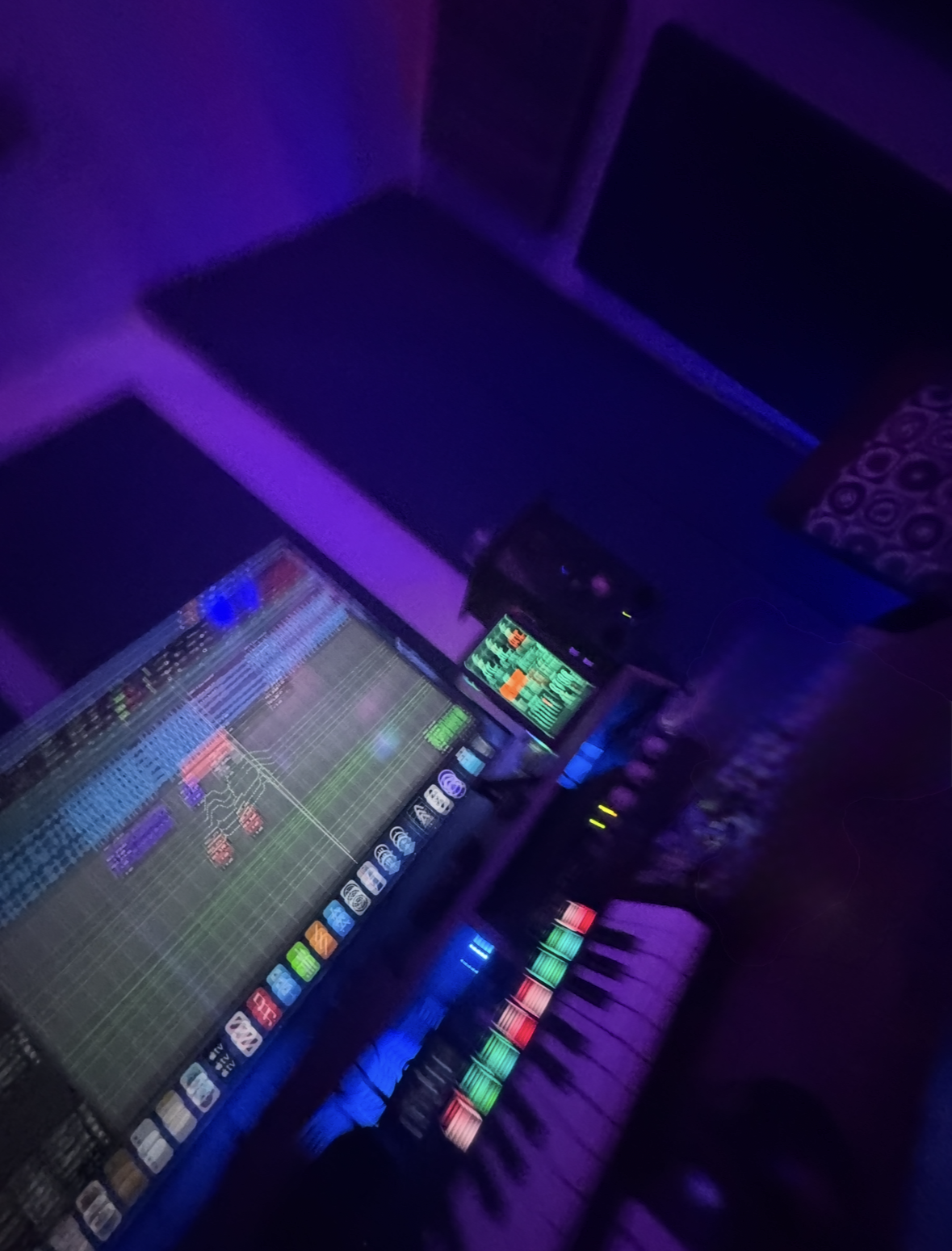 A DJ setup in a dimly lit room with purple and blue lighting, featuring a laptop, DJ mixers, and a keyboard with colorful buttons. There are speakers and a small LED screen displaying sound levels.