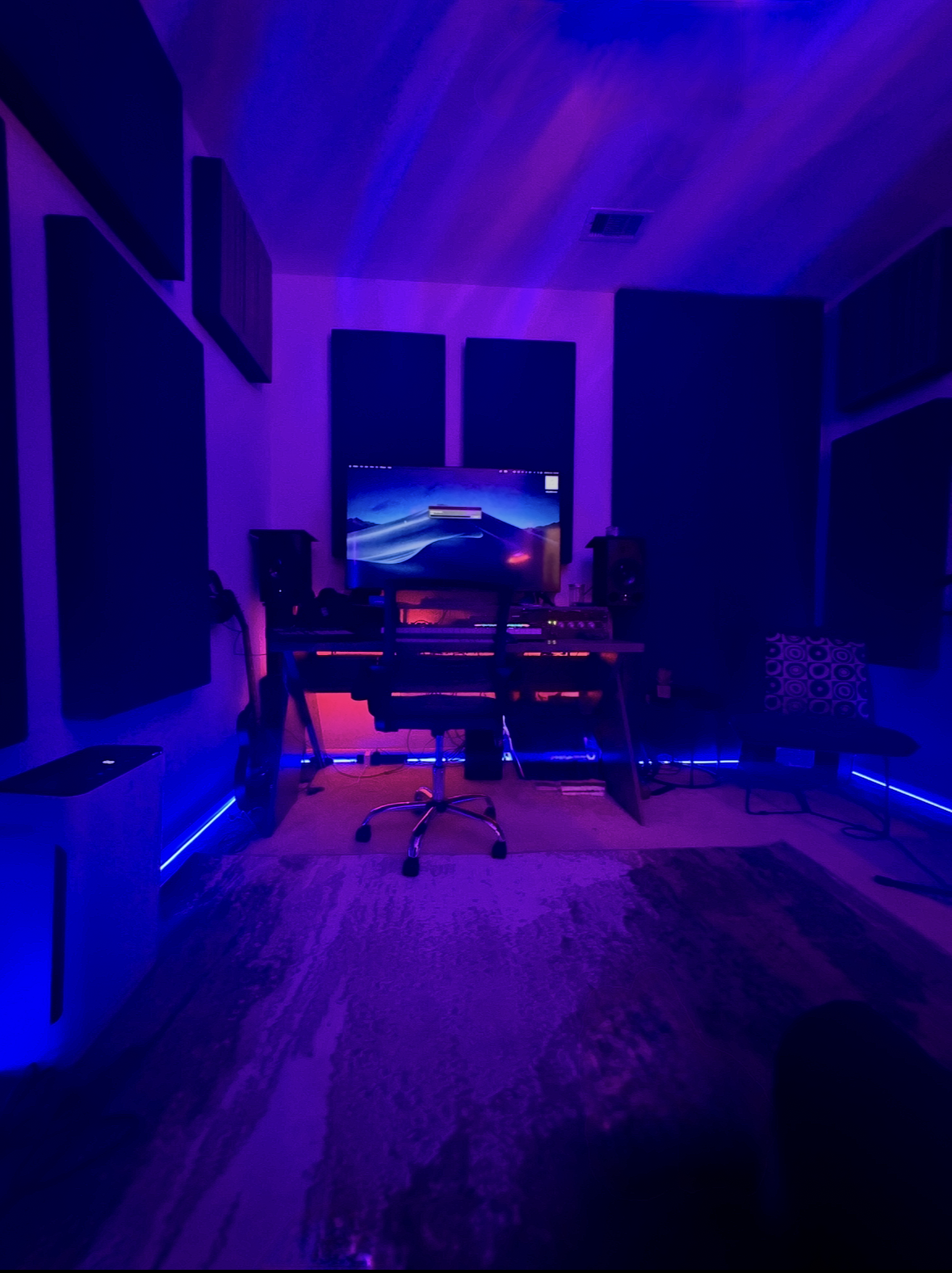 A music or recording studio with purple and blue LED lighting, a computer monitor on a desk with audio equipment, speakers, and a chair.