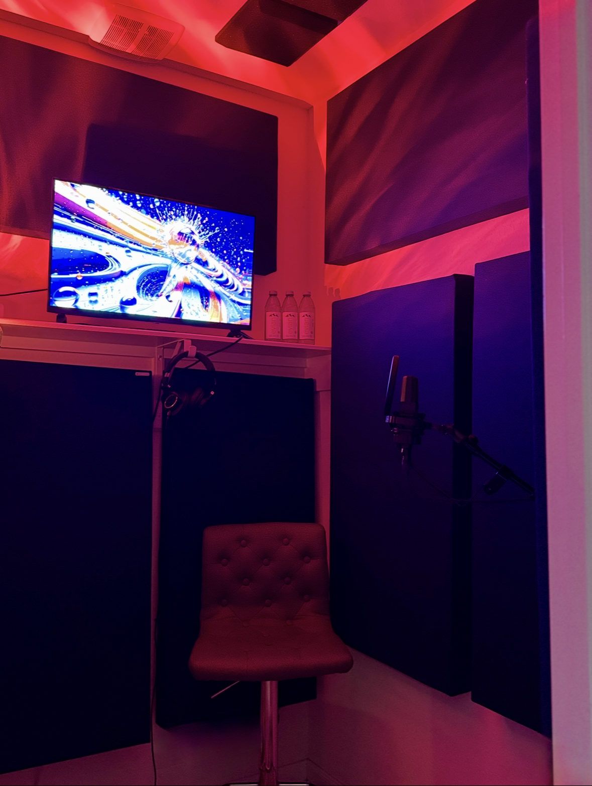 Recording studio with a monitor displaying colorful visuals, black soundproof panels on the walls, a microphone on a stand, a red cushioned stool, and three water bottles on a shelf in a low ambient red and purple lighting environment.