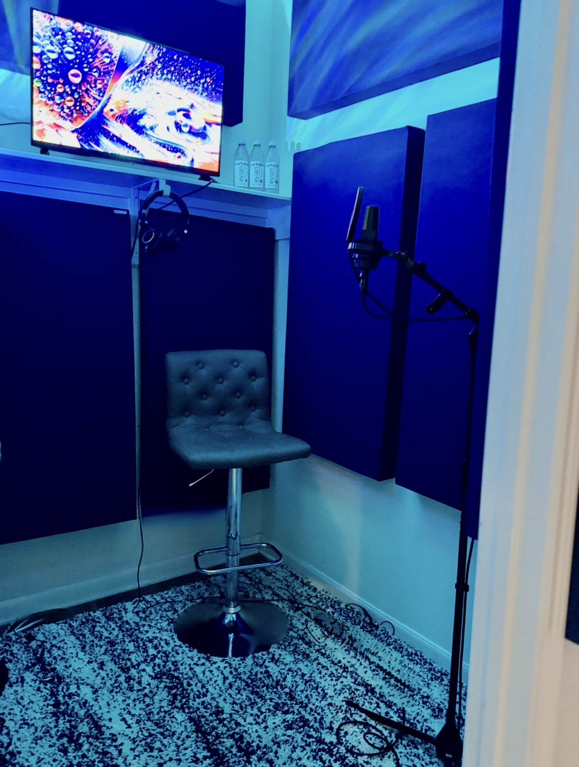 Music recording isolation booth with a chair, AKG C414 micophone, headphones, monitor, and Evian water bottles, illuminated by blue lighting.