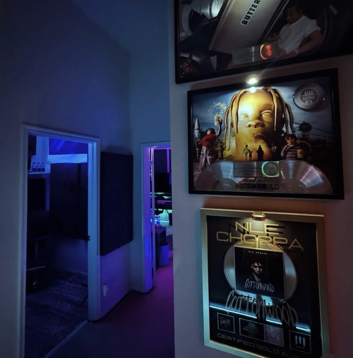 Wall with framed music awards and posters, dimly lit with colorful ambient lighting, revealing parts of an adjoining room with purple and blue lighting.