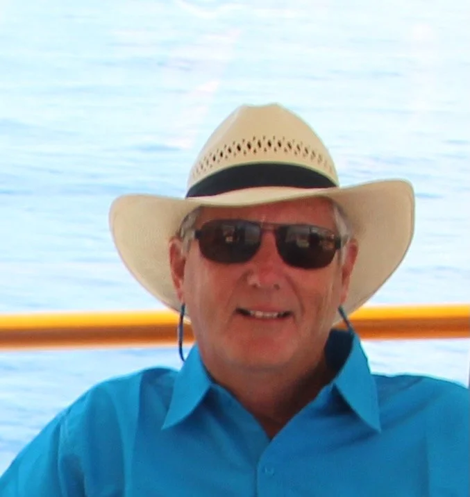 Author Anthony McGruther, wearing a beige sun hat, dark sunglasses, a blue shirt, and earplugs, smiling on a boat with water in the background.