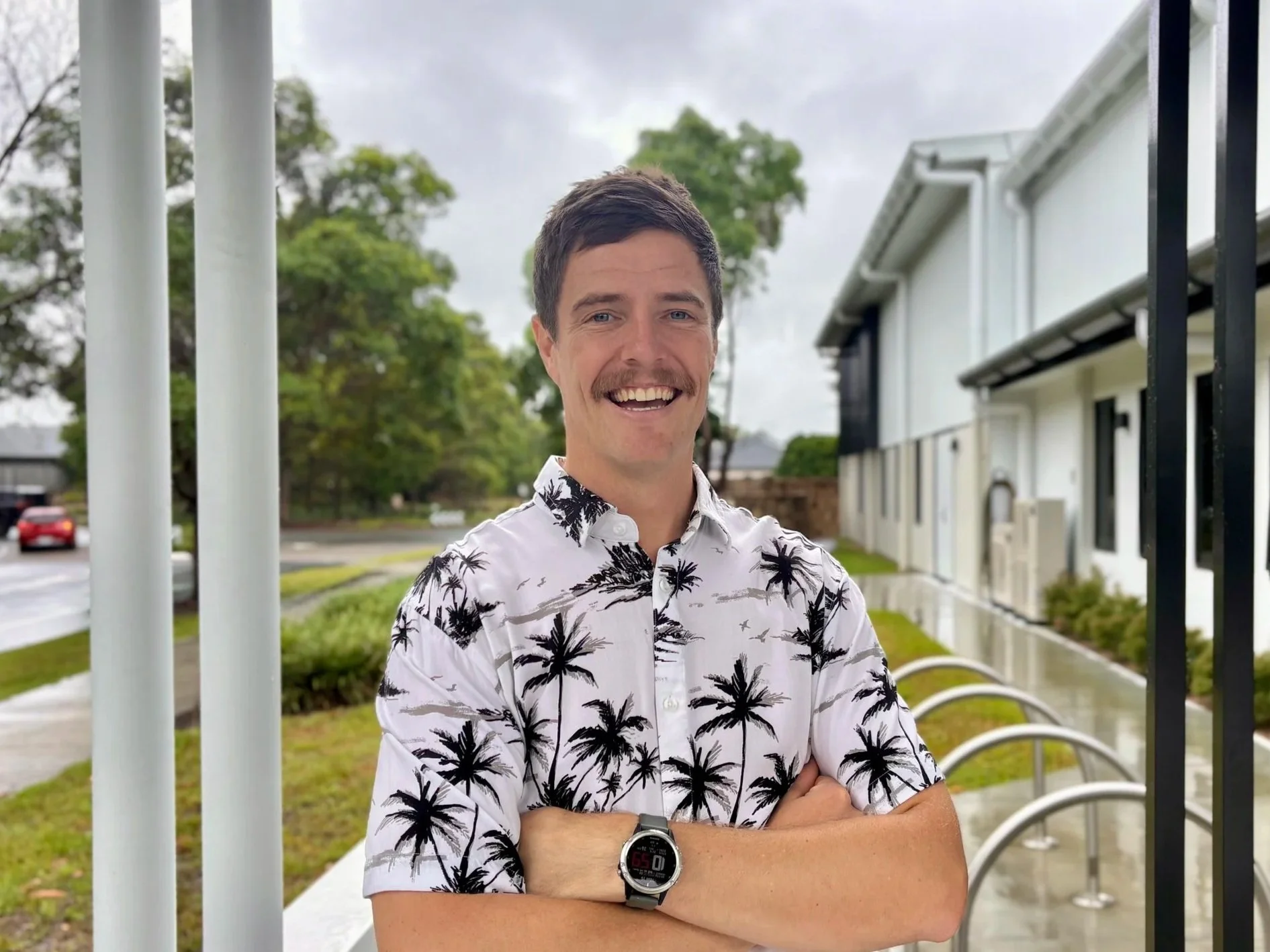 A smiling man with a mustache wearing a white shirt with black palm tree patterns, standing outdoors with houses and trees in the background on a cloudy day.