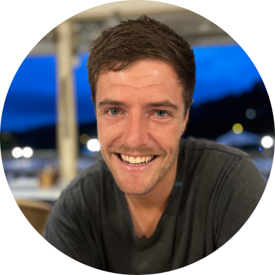 A smiling man with short brown hair and a dark T-shirt, sitting outdoors during evening with a blurred background of lights and sky.