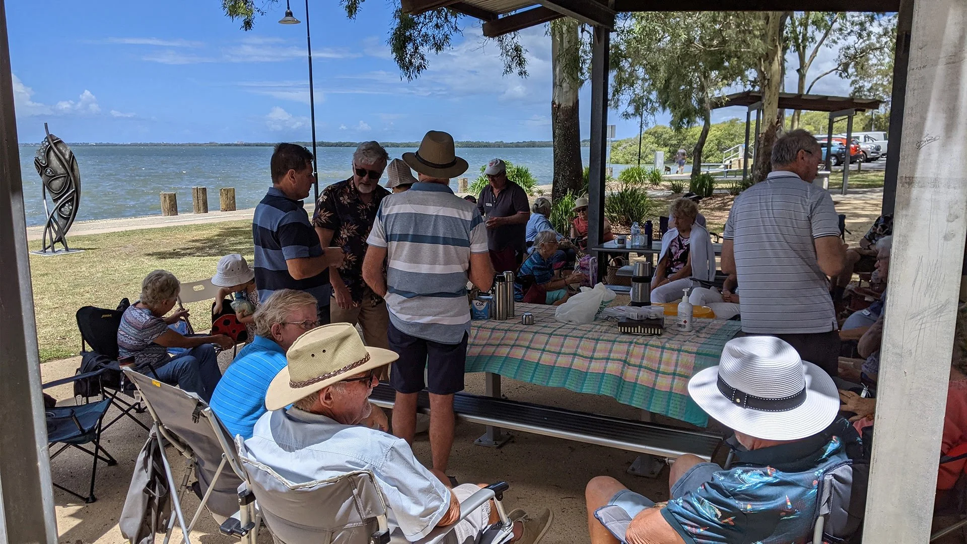 A group of people gathered under a tent near a body of water, some sitting and some standing, with trees and a walkway in the background.