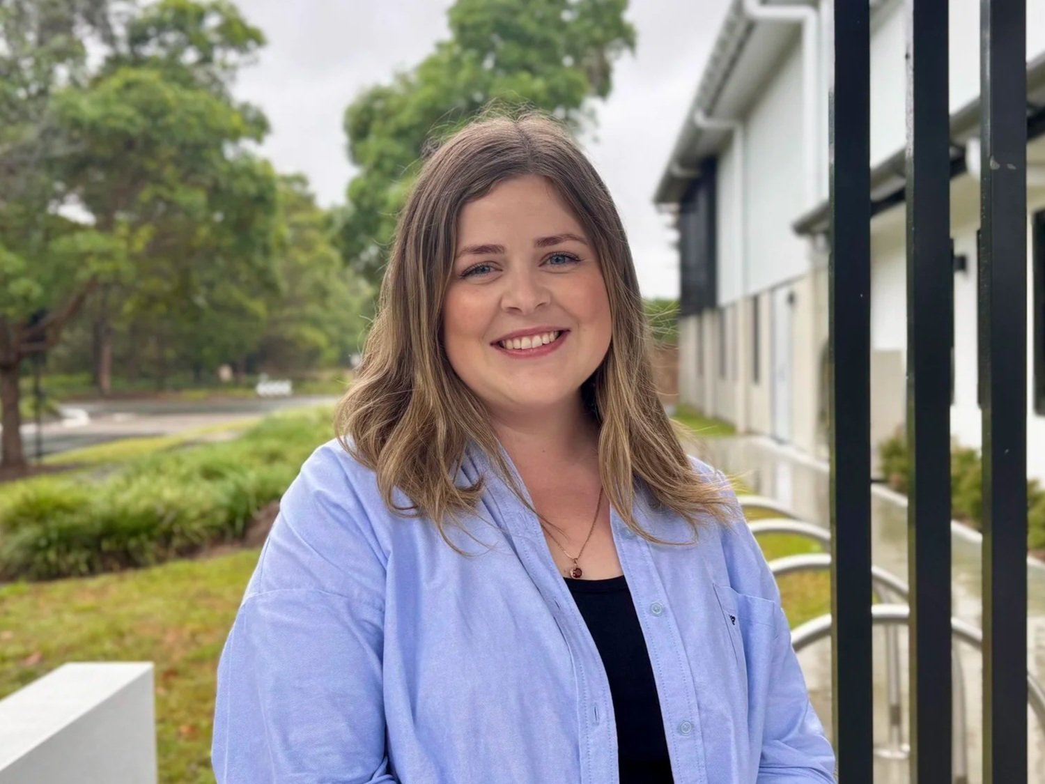 A young woman with light brown, wavy hair smiling at the camera, wearing a green and white striped shirt and a gold necklace.