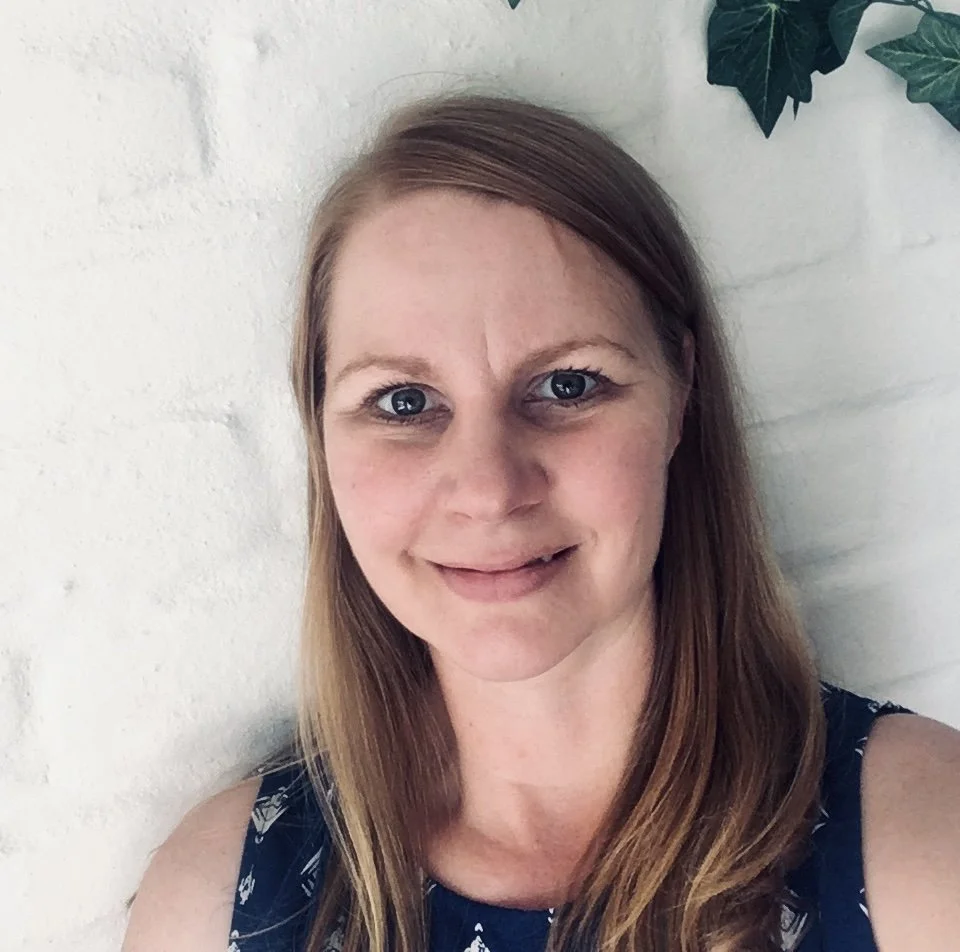 A woman with long, light brown hair, blue eyes, and fair skin, smiling and wearing a sleeveless dark blue top with a pattern, standing against a white brick wall with some green leaves in the upper right corner.