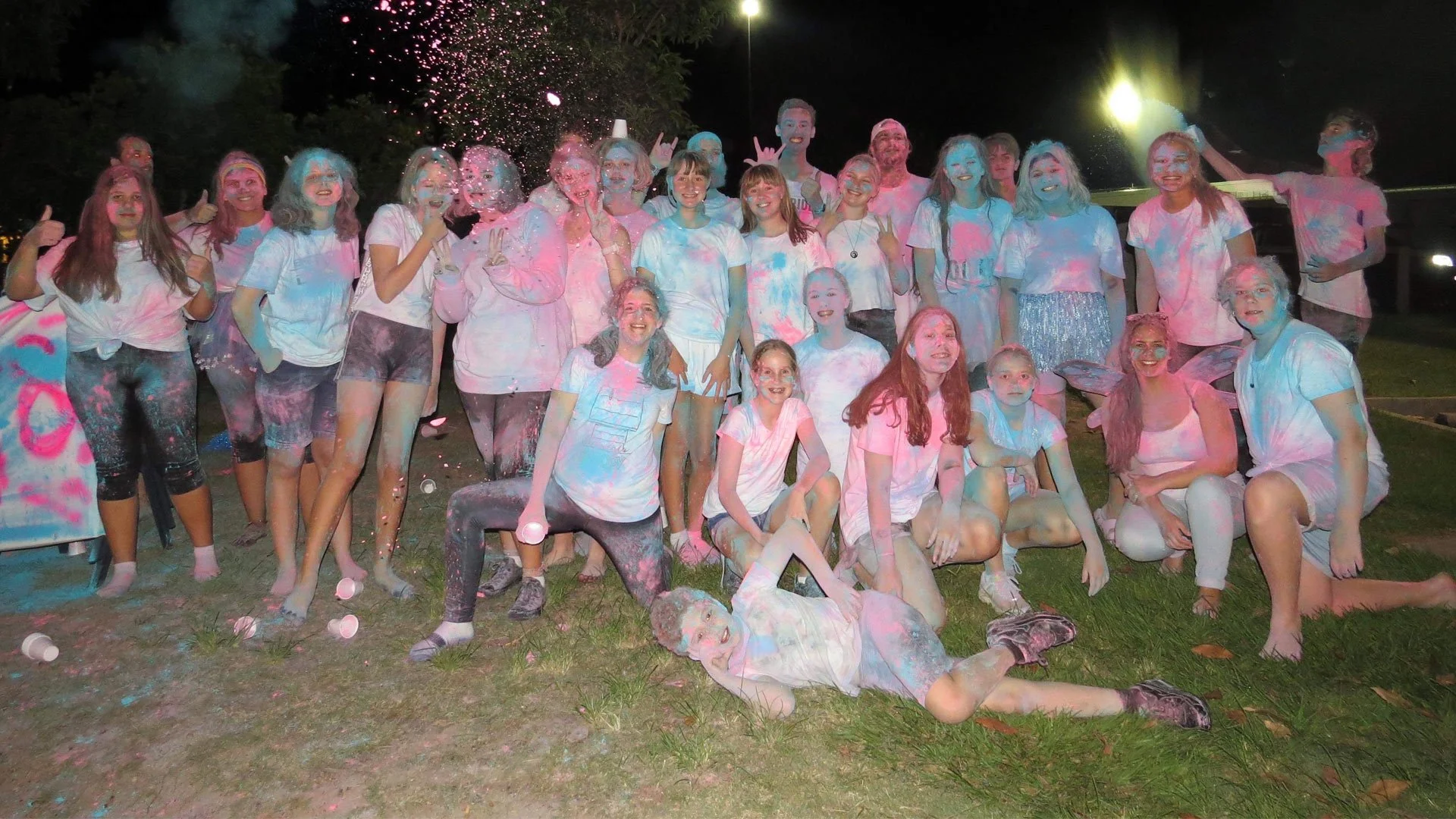 A group of young people at night celebrating during a color powder event, with some throwing powder and others covered in pink, blue, and white powder, smiling, and posing outdoors.