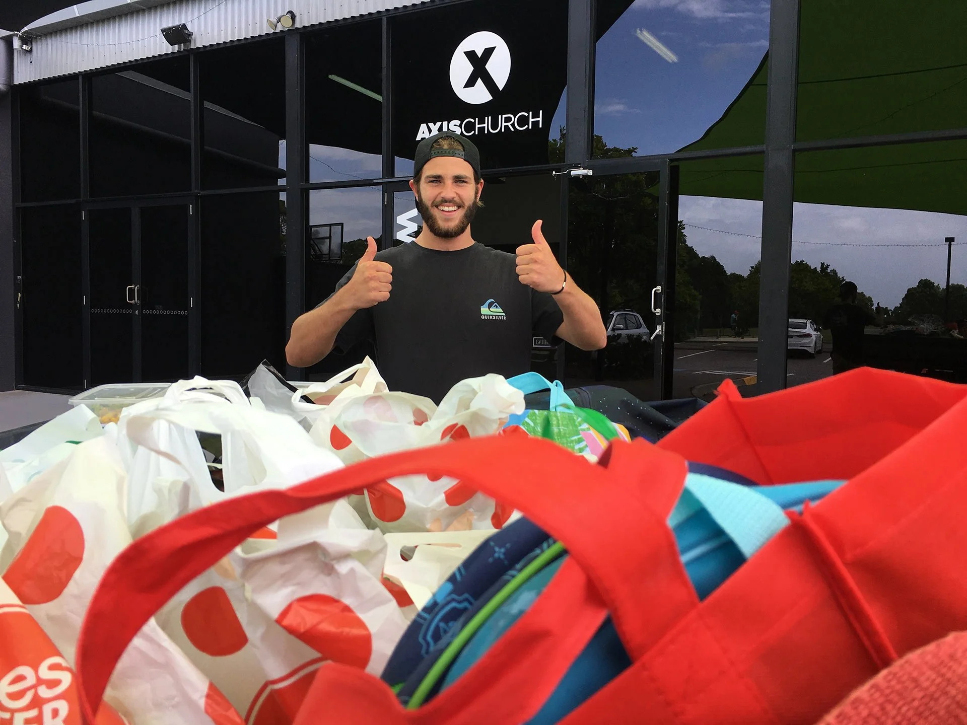 A man giving a thumbs up outside an AXISCHURCH building, with colorful bags and items on a table in the foreground.