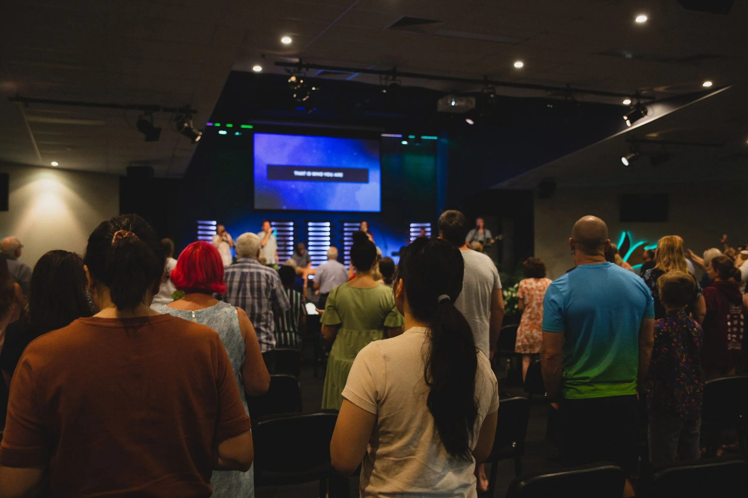 A crowd of people attending a worship service or concert in a dimly lit auditorium. The congregation is standing, facing a stage where musicians and singers are performing. The stage has a large screen displaying text and stage lights.