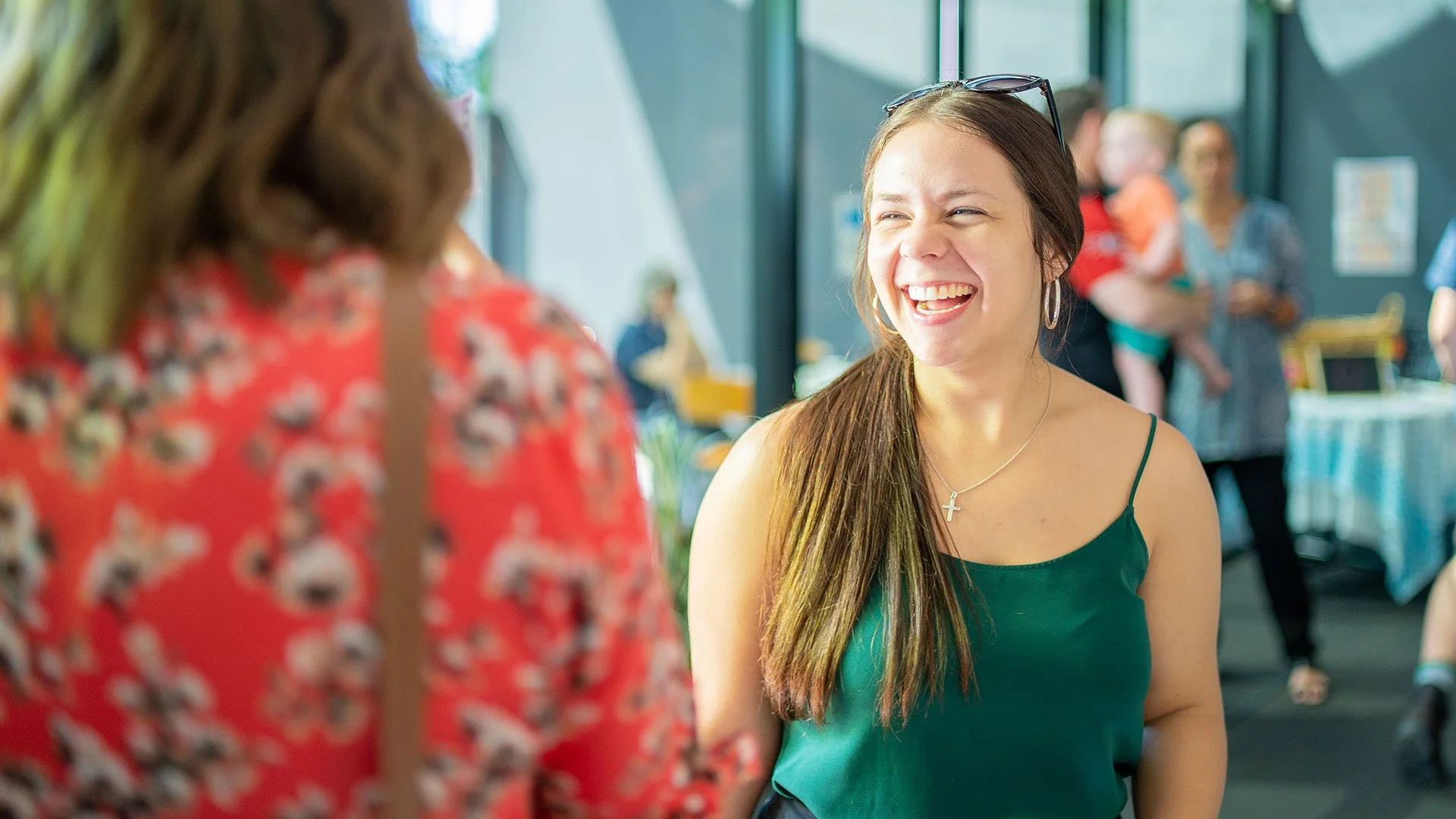 A woman with long brown hair smiling and laughing while talking to someone at an indoor social gathering. She is wearing a green sleeveless top, hoop earrings, and a cross necklace. Other people are visible in the background.